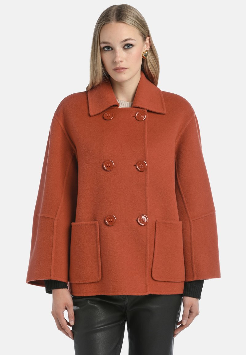 Manteau double boutonnage couleur rouille avec des revers surdimensionnés, deux poches avant et des manches larges. Fabriqué dans un tissu texturé à la finition douce.