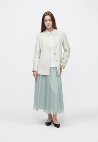 Blazer de linho branco com botões dourados sobre uma camisa branca, combinado com uma saia de tule verde claro e sapatilhas pretas.