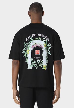 Mand iført sort T-shirt med teksten "PAS DE MONACO" og grafisk blomsterbue med grønne blade, gule citroner og lilla blomster på bagsiden.