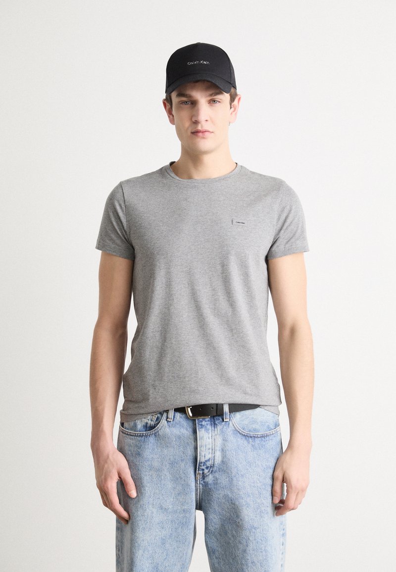 Γκρι βαμβακερό t-shirt με στρογγυλή λαιμόκοψη και κοντά μανίκια, που φέρει μία μικρή μαύρη ετικέτα. Συνδυάζεται με ανοιχτό μπλε τζιν παντελόνι και μαύρο καπέλο.