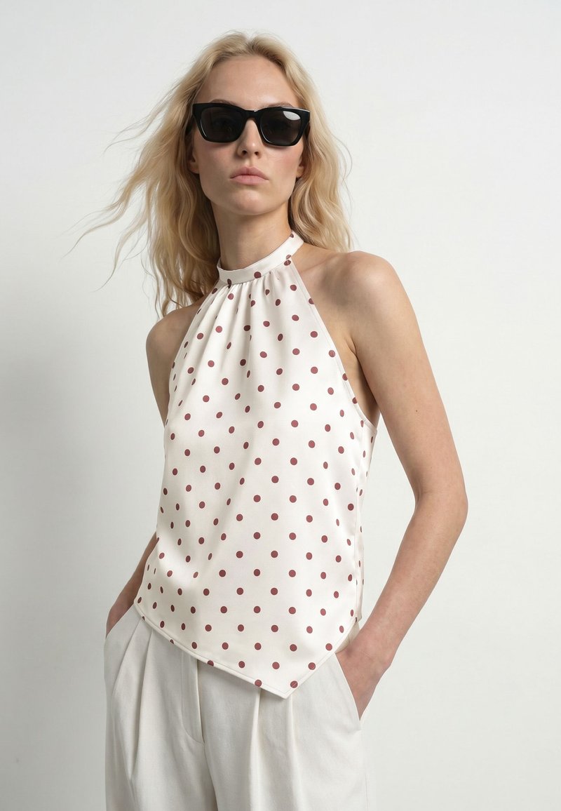 Femme blonde portant des lunettes de soleil noires, un haut dos nu blanc à pois rouges et un pantalon blanc, debout devant un fond clair uni.