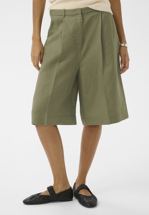 Kobieta w oliwkowozielonych, szerokich szortach do kolan i czarnych balerinach z marszczeniami, stojąca na białym tle.