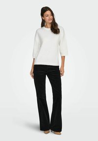 Pull en maille blanc à manches courtes avec un col rond, associé à un pantalon évasé en velours côtelé noir taille haute et des talons beiges.