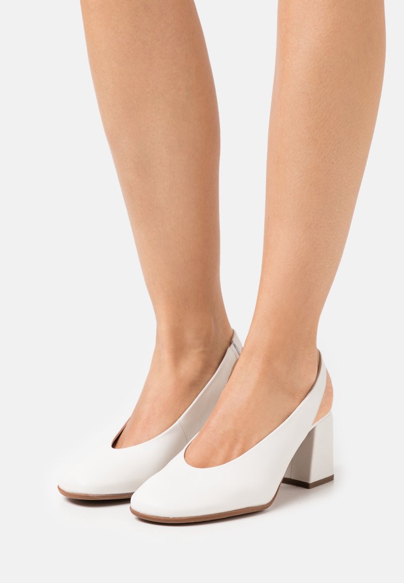 Žena v bílých kožených slingback botách na blokovém podpatku se zakulacenými špičkami na jemném světlém pozadí.