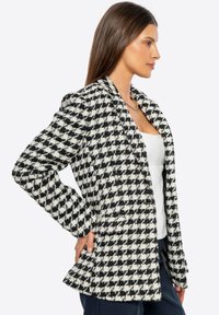 Blazer de patrón houndstooth en blanco y negro con una tela texturizada, corte ajustado, solapas con muesca y dos bolsillos frontales.