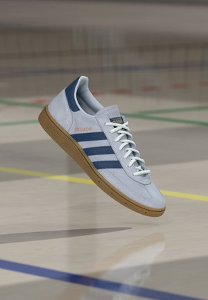 Vaaleanharmaa Adidas Spezial -lenkkari, jossa tummansinisiä raitoja, valkoiset kengännauhat ja kumipohja, leijuu värillisten linjojen päällä kuntosalisalilla.