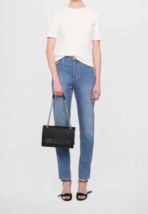 Persoon draagt een wit kort mouwtopje, blauwe spijkerbroek, zwarte sandalen met hoge hakken en houdt een zwarte Love Moschino handtas met kettingriem vast.