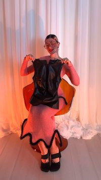 Haut noir sans manches en matériau brillant présenté sur une longue robe rose côtelée avec un bas ondulé orné de fourrure et des chaussures noires.