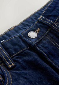 Zbliżenie na granatowe dżinsy z żółtym przeszyciem i srebrnym guzikiem z napisem. Tkanina jest lekko fakturowana.