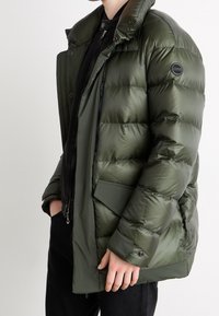 Chaqueta acolchada verde oliva con cierre frontal, bolsillos laterales y un cuello alto; presenta una textura suave y brillante con un parche de logo.