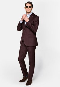 Bordowy szeleszczący garnitur o slim fit, z marynarką na jeden guzik i dopasowanymi spodniami. Jasnoniebieska koszula pod spodem, brązowe zamszowe buty, stylowe okulary przeciwsłoneczne.