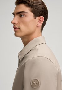 Beżowa kurtka z wyprofilowanym kołnierzem, zapięciem na zamek i okrągłą naszywką z logo na lewym rękawie. Gładka faktura z dopasowanym krojem.