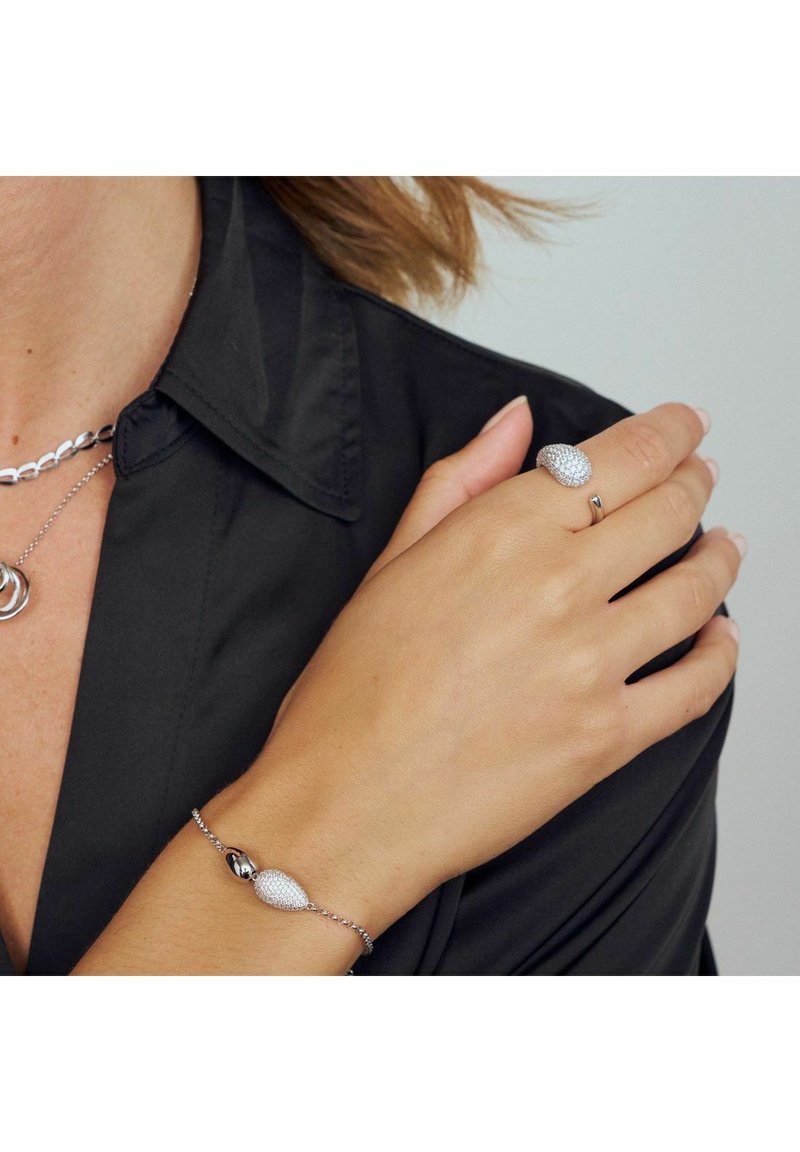 En kvinna i en svart skjorta visar ett silverarmband och två silverringar på sin vänstra hand mot en neutral bakgrund.