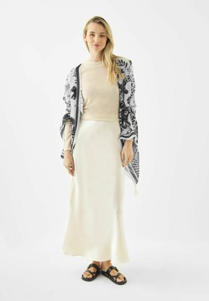 Femme portant un haut à manches longues beige, une jupe en satin crème, un châle floral noir et blanc, et des sandales noires, debout devant un fond blanc uni.