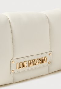 Balta mākslīgā ādas rokassoma ar kvadrātu dizainu, kurai ir zelta toņa metāla plāksne ar uzrakstu "LOVE MOSCHINO" un dekoratīvā izšūšana.