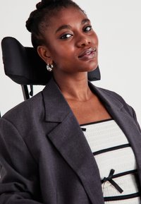 Γκρι σακάκι που έχει ραφτεί προσεκτικά πάνω από λευκό μπλουζάκι με μαύρες ρίγες και λεπτομέρεια με φιόγκο. Το μοντέλο έχει πλεξούδες και φορά ασημένια σκουλαρίκια κρίκους, καθισμένο σε αναπηρικό αμαξίδιο.