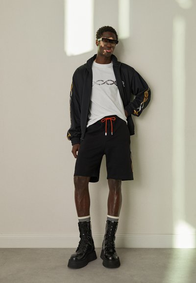 Μαύρο μπουφάν τύπου bomber με πορτοκαλί λεπτομέρειες, λευκό μπλουζάκι με γραφικά, μαύρες βερμούδες με πορτοκαλί κορδόνι και μαύρες πλατφόρμες μποτάκια με ριμπ ά socks.