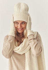 Beanie a maglia color avorio con risvolto a coste, guanti a maglia coordinati con texture e una sciarpa morbida. Tutti i pezzi presentano delicati motivi a treccia.