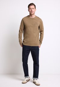 Světle hnědá košile s dlouhým rukávem a potiskem na přední straně, spárovaná s tmavě modrými džíny a bílými teniskami, s hnědými akcenty a ohrnutými rukávy.