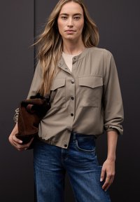 Veste boutonnée marron clair avec deux poches poitrine, manches froncées et texture lisse, portée avec un jean bleu. Sac marron tenu à la main.