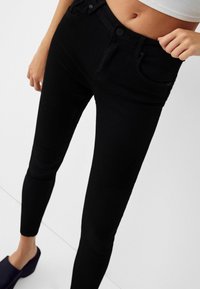 Mujer vestida con jeans ajustados de cintura alta negros y zapatos negros tipo slip-on, ajustándose la cintura contra un fondo blanco.
