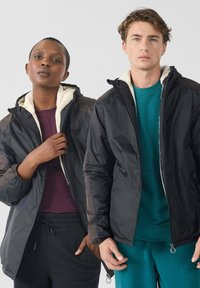 Twee modellen dragen zwarte hoodies met witte fleece voering; één stelt de jas bij, beiden staan tegen een effen achtergrond.