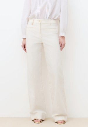Witte wijde broek gedragen met een witte blouse met lange mouwen en beige sandalen, gezien van taille tot onder tegen een witte achtergrond.