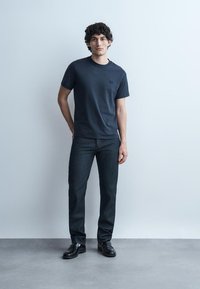 Mladi moški s kodrastimi lasmi, oblečen v temno mornarsko modro majico, temne kavbojke in črne usnjene čevlje, stoji ob svetlo sivi steni.