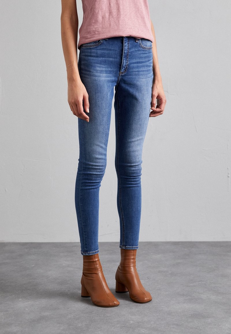 Femme portant un jean slim bleu, des bottines marron à talons carrés, et une chemise rose clair, debout sur un sol gris devant un mur gris.