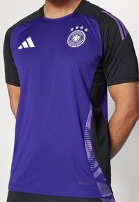 Lila sporttröja med svarta ärmar. Har rund hals, Adidas-logotyp och ett femstjärnigt emblem på bröstet. Texturerad sidopaneldesign.