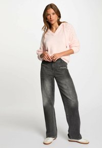 Maglione rosa chiaro, morbido e lavorato a maglia, con scollo a V e cappuccio, abbinato a jeans grigi a vita alta con gambe larghe e tasche decorative. Sneakers bianche.