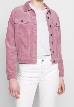 Personne portant une veste en velours côtelé rose par-dessus un t-shirt blanc et un pantalon blanc taille haute, debout devant un fond uni.
