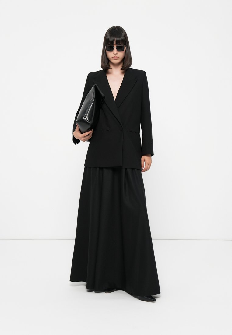 Blazer noir ajusté avec des revers nets, porté sur une longue jupe noire. Une grande pochette noire et des lunettes de soleil noires élégantes complètent le look.