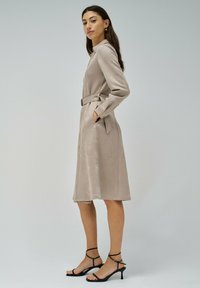 Vestido beige con cinturón, cuello y manga larga, que cuenta con bolsillos laterales y una textura suave, combinado con tacones negros de tiras.