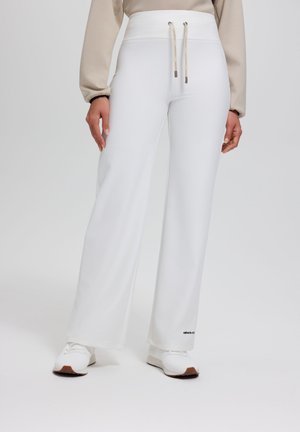 Frau trägt weiße, weit geschnittene Hose mit Kordelzug in der Taille und weiße Turnschuhe, steht vor einfachem Hintergrund.