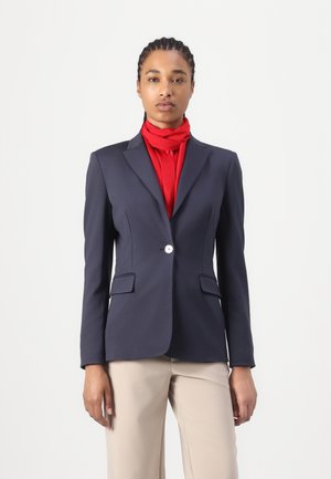 Blazer marine à la coupe cintrée, fermeture à un bouton et deux poches sur le devant. Porté avec une écharpe rouge et associé à un pantalon beige clair.