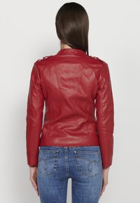 Red leather jacket with fitted design, stitch detailing, and shoulder accents. Paired with blue jeans featuring decorative back pockets.