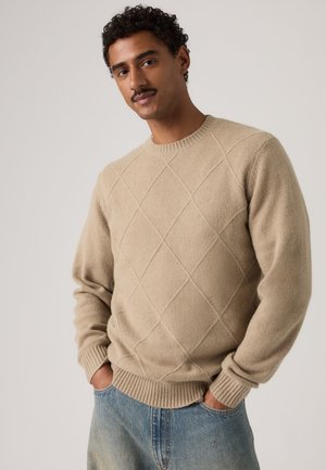 Hombre con pelo rizado negro y bigote que lleva un suéter beige con patrón de diamantes y vaqueros azules con las manos en los bolsillos sobre un fondo liso.