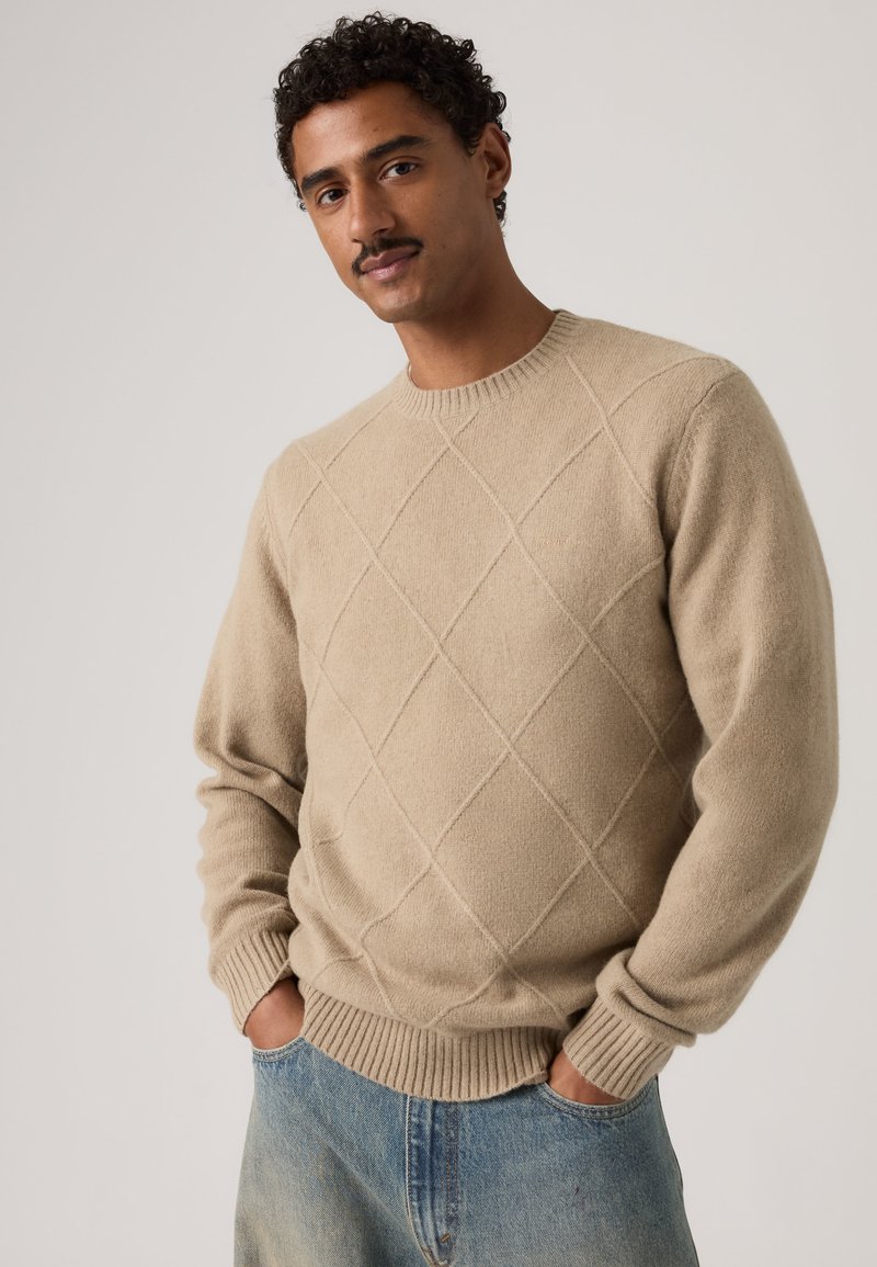 Homme aux cheveux noirs bouclés et moustache portant un pull beige à motif losange et un jean bleu, mains dans les poches, sur fond uni.