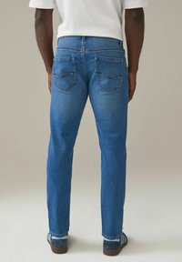 Hombre vestido con pantalones vaqueros rectos de color azul medio, camiseta blanca, calcetines azul claros y zapatillas deportivas azul marino, de pie mirando hacia atrás sobre un fondo liso.