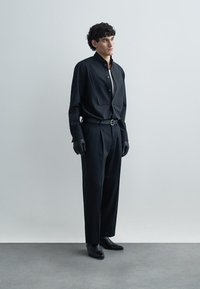 Chemise noire à manches longues avec col mao, associée à un pantalon noir taille haute et des gants noirs ; design épuré et minimaliste.