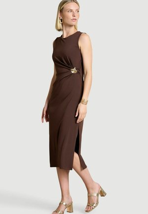 Frau trägt ärmelloses braunes Midikleid mit Seitenschlitz und goldener Schnallendetail, kombiniert mit goldenen Sandalen mit Blockabsatz und Schmuck.