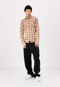 Brun og beige rutete langermet skjorte med knappeåpning, kombinert med baggy svarte jeans og hvite joggesko. Modellen har på seg en svart lue.