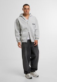 Gray zip-up hoodie with a front pocket and logo, paired with baggy black denim pants and multi-colored sneakers. Smooth texture.