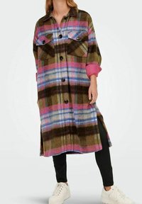 Woman wearing a long plaid coat with pink, blue, and olive stripes, black pants, rolled sleeves, and white platform sneakers.