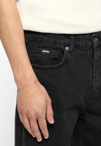 Δεξί χέρι με λεπτό ασημένιο βραχιόλι που ακουμπάει δίπλα στην τσέπη μαύρου τζιν τζιν και λευκό πλεκτό πάνω μέρος ορατό πάνω από τη ζώνη.