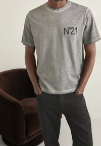 Szara bawełniana koszulka z krótkim rękawem i okrągłym dekoltem, z małym czarnym nadrukiem "N°21" na piersi, zestawiona z czarnymi spodniami, na tle brązowego welurowego fotela.