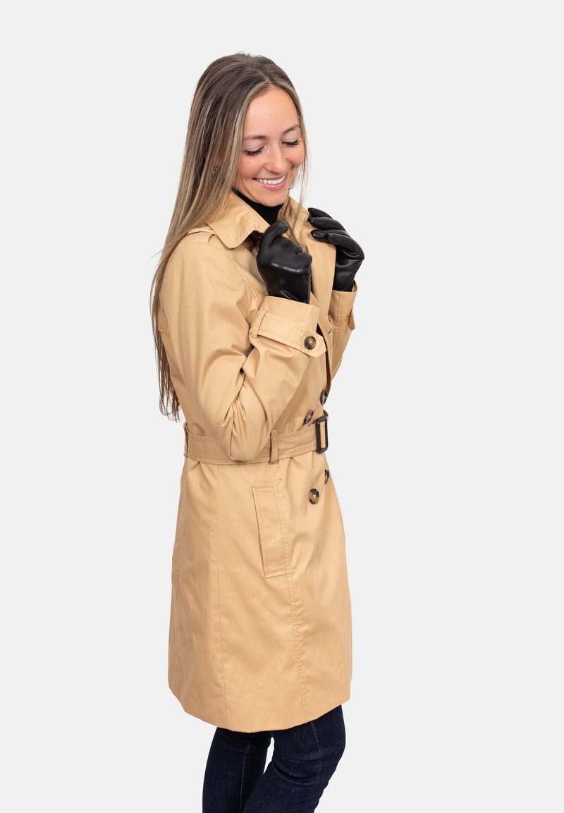 Beiger Trenchcoat mit tailliertem Gürtel, Knopfdetails und zwei Seitentaschen. Kombiniert mit schwarzen Handschuhen und dunklen Jeans.