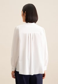 Blusa bianca con vestibilità ampia, maniche lunghe arricciate, orlo arrotondato e dettagli di pieghe sottili sul retro. Tessuto liscio con una trama morbida.