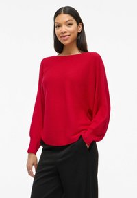 Pull surdimensionné en tricot côtelé rouge avec des manches longues, doté d'un large col et d'une coupe décontractée, associé à un pantalon noir.